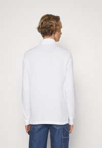 Hvit langermet polo-skjorte med krage, glatt tekstur og sidesplitter. Bakvinkelen viser et rent design og en tettsittende silhuett.