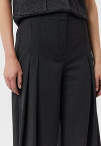 Spodnie o szerokich nogawkach wykonane z ciemnoszarej tkaniny, charakteryzujące się subtelną fakturą, plisami z przodu i gładkim pasem, pokazane z dłonią opartą na boku.