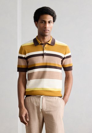 Man staat en draagt een polo met korte mouwen met horizontale strepen in bruin, mosterdgeel, beige en wit en lichtbeige broek met trekkoord.