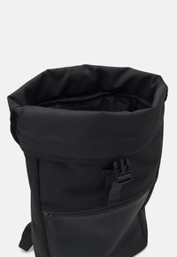 Grand sac à dos noir à rabat avec une poche zippée à l'avant et une fermeture à boucle en plastique sur un fond uni.