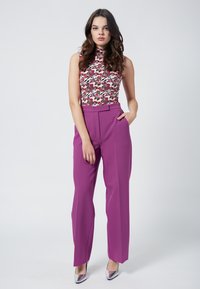 Figurbetontes, ärmelloses Floral-Top, kombiniert mit hochgeschnittenen, lila Hosen. Das Model trägt metallic silberne High Heels. Einfarbiger Hintergrund.