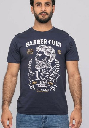Homme portant un t-shirt bleu marine avec un design vintage de barbier, représentant un homme barbu tenant des tondeuses et le texte « Barber Cult » et « Old Glory ».