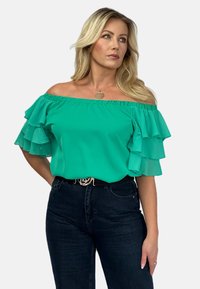 Turkusowa bluzka typu off-shoulder z warstwowymi falbanami na rękawach. Gładki, lekki materiał zestawiony z ciemnymi jeansami z wysokim stanem i paskiem.