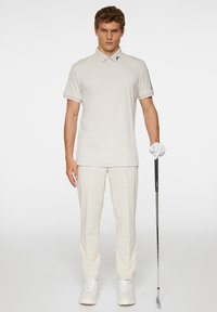 Ljusbeige pikétröja med krage och korta ärmar, i kombination med krämfärgade byxor och vita skor, som håller en golfklubba.