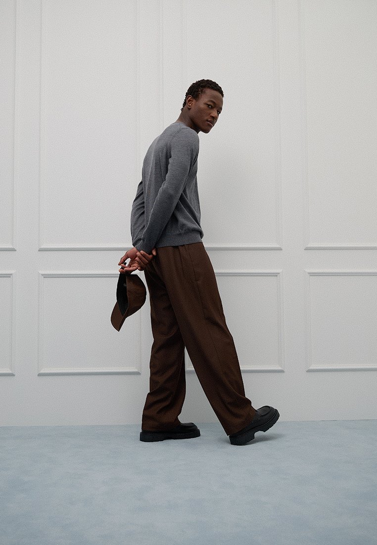 Maglione grigio, pantaloni marroni e scarpe nere. L'outfit presenta una vestibilità comoda e una texture liscia, abbinato a un cappellino indossato al contrario.