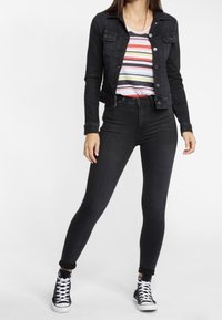 Giacca in denim nera su una camicia a righe con linee rosa, gialle e grigie. Jeans skinny neri e scarpe alte nere.