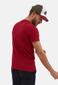 Camiseta roja de manga corta con cuello redondo y un corte holgado, combinada con pantalones oscuros. Presenta un sutil logo en la parte posterior.