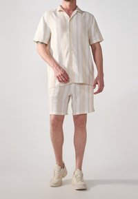 Camisa de manga corta, abotonada, y pantalones cortos a juego en rayas verticales crema y beige, confeccionados en tejido ligero. Se añaden zapatillas casuales.