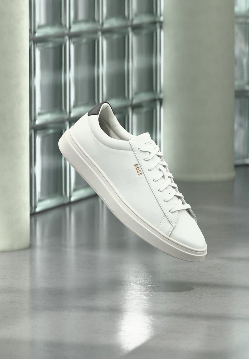 Baskets basses en cuir blanc avec lacets, languette de talon noire et logo « BOSS », flottant à l'intérieur près d'un mur en blocs de verre et d'un sol poli.