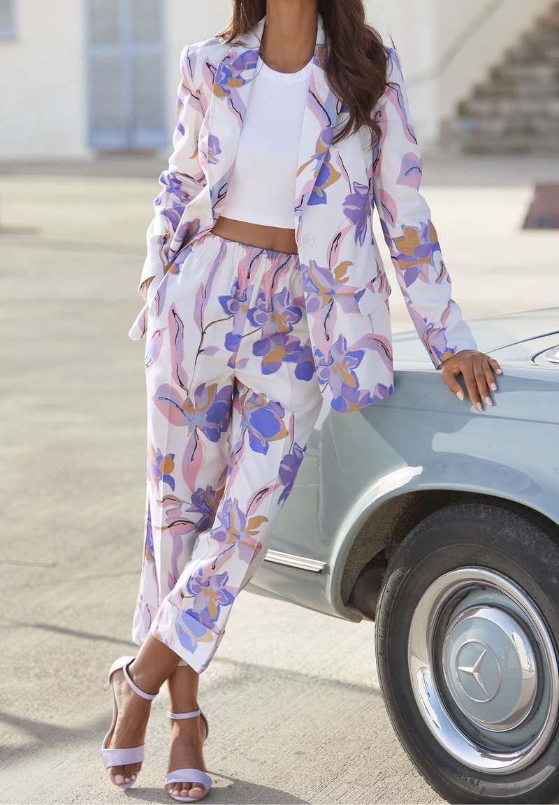 Femme portant un blazer et un pantalon à fleurs pastel, un crop top blanc et des talons, appuyée sur une voiture Mercedes vintage bleu clair en extérieur.