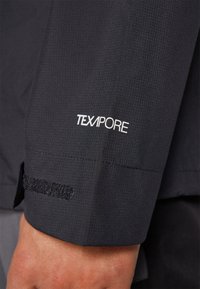 Gros plan sur la manche d'une veste noire avec le logo blanc "TEXAPORE" et une sangle Velcro ajustable au-dessus du poignet sur le bras d'une personne.