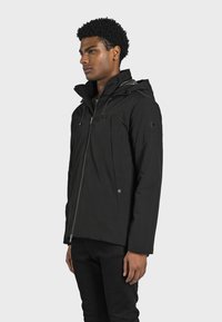Sort vandtæt jakke med hætte, lynlås foran, to sidelommer og justerbare snore. Glat tekstur og minimalistisk design.