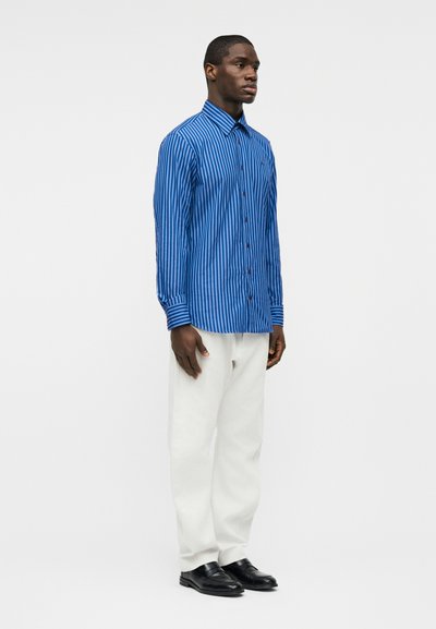 Άνδρας όρθιος, φορώντας μπλε ριγέ πουκάμισο με κουμπιά, λευκό παντελόνι και μαύρα παπούτσια, μπροστά σε απλό ανοιχτό φόντο.