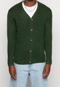 Donkergroene gebreide cardigan met een V-hals, voorzien van zeven bruine knopen en geribbelde manchetten en zoom. Draagt over een wit shirt en lichtblauwe spijkerbroek.