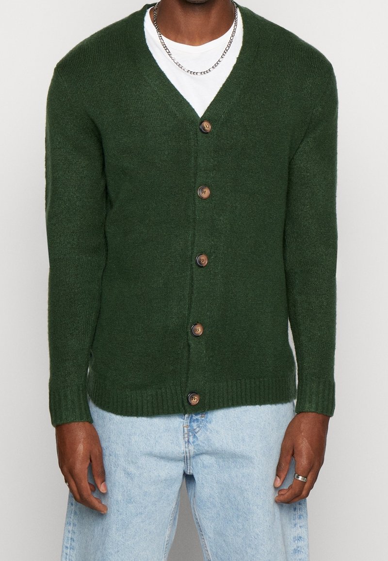 Donkergroene gebreide cardigan met een V-hals, voorzien van zeven bruine knopen en geribbelde manchetten en zoom. Draagt over een wit shirt en lichtblauwe spijkerbroek.