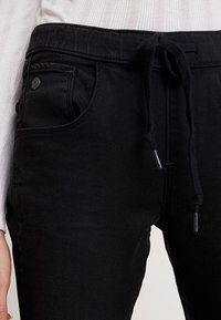 Svarta joggerbyxor i bomull med dragsko i midjan, sidofickor och mjuk textur. Har en metallknappdetalj på framfickan.