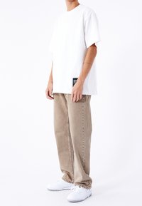 Camiseta de algodón blanca de ajuste holgado, combinada con pantalones vaqueros beige de pierna recta. Zapatillas blancas completan el conjunto. Detalle de etiqueta negra en la camisa.