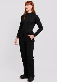 Schwarzes Langarmoberteil und schwarze Hose mit Taschen, im figurbetonten Design und mit glatter Textur. Das Modell steht in einer neutralen Pose.