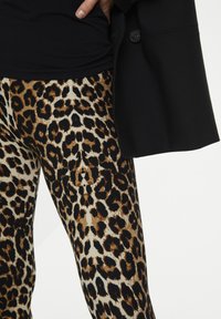 Leggings s uzorkom leoparda imaju kombinaciju boja prirodne braon, smeđe i crne, izrađene su od rastezljivog materijala. Crni top i kaput su delimično vidljivi.