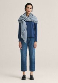 Sininen villa-neule, vaaleansininen fringed-huivi, korkeavyötäröiset siniset farkut ja mustat loafertossut. Neutraali tausta.