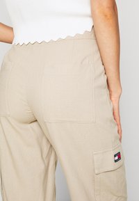 Beige cargo pants made of lightweight fabric, featuring two large back pockets and a small logo patch on the side. Textured with a smooth finish.