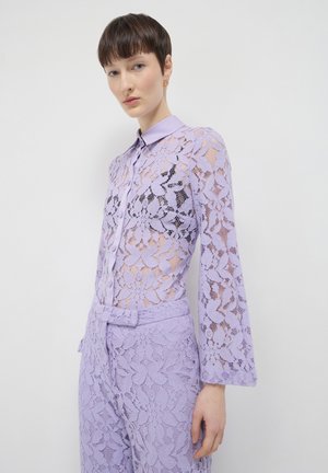 Woman wearing a sheer lavender floral lace blouse with collar and matching high-waisted lace pants against a plain white background.