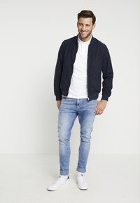 Giubbotto bomber navy con una texture liscia, con zip frontale, indossato sopra una maglietta polo bianca, abbinato a jeans tapered azzurri chiari e sneaker bianche.