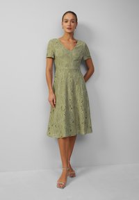 Robe en dentelle verte à manches courtes, corsage ajusté et jupe évasée. Décolleté en V et motif floral. Portée avec des sandales à lanières argentées.
