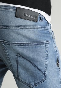 Lichtblauwe denim jeans met een contrasterend zwart leren label met de tekst "CHASIN'." Bevat schuine achterzakken met zichtbare stikseldetails.