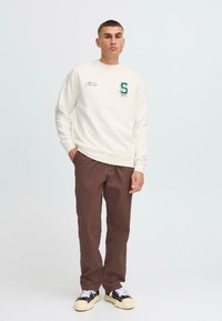 Tricou alb cu broderie verde "S" și text pe piept, purtat împreună cu pantaloni maro și adidași alb-negru.