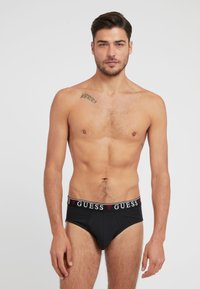 Schwarze Baumwollslips mit einem elastischen Bund mit Markenschriftzug, der weißen und roten "GUESS"-Schriftzug zeigt, entworfen für eine enganliegende Passform.
