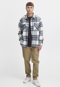 Karo-Flanellhemd in Weiß, Blau und Schwarz, mit einer Tasche und geknöpftem Front, getragen über einem marineblauen T-Shirt, kombiniert mit beigen Hosen und weißen Sneakers.