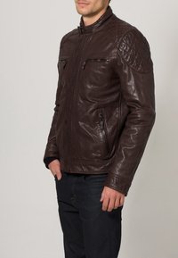 Hnědá kožená bunda s prošívanými akcenty na ramenou, dvěma předními kapsami na zip a límcem na patentky. Hladká textura a přiléhavý střih.