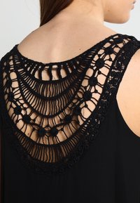 Czarny top bez rękawów z misternym czarnym koronkowym wykończeniem na plecach, z kwiatowym i zakrzywionym wzorem, podkreślającym teksturę i elementy designu.