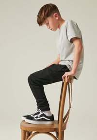 Tricou gri din bumbac, pantaloni negri de trening cu buzunare laterale și adidași Vans negri cu tălpi albe, așezat pe un scaun din lemn.