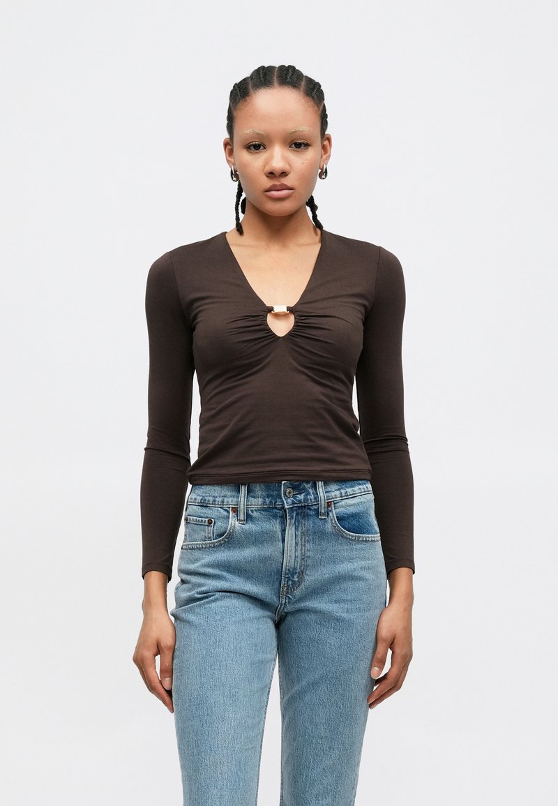 Chemise marron à manches longues avec un col en V et un détail froncé à l'avant, ornée d'un accent métallique au niveau de l'encolure. Associée à un jean bleu clair.