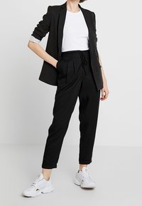 Černý blejzr s ohrnutými rukávy, bílý tričko, černé kalhoty na stahování a bílé sportovní boty. Jednoduchý design, hladké textury a přiléhavý střih.