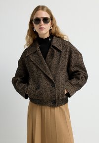 Bruine herringbone-geborduurde jas met een dubbelzijdig ontwerp, grote revers en zwarte knopen, gecombineerd met een zwarte coltrui en een geplooide rok.