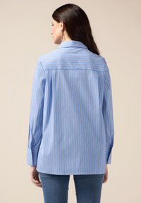 Blusa a rayas azul con rayas verticales en blanco y naranja. Presenta un cuello, mangas largas y un corte relajado. Textura de tela suave.