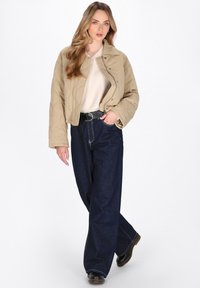 Beige quiltet jakke med en cropped design, parret med højtaljede mørkeblå vide jeans. Modellen har sorte ankelstøvler på.