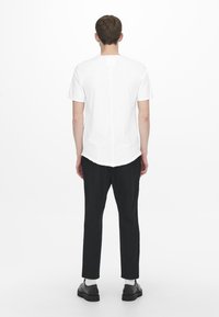 Witte t-shirt met korte mouwen en een rechte achterkant, gedragen met zwarte broek en zwarte schoenen, wat een eenvoudig, minimalistisch ontwerp laat zien.