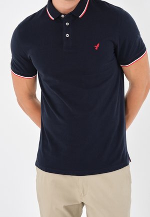 Navy polo shirt with a red bird logo, white and red striped collar and sleeve hems, features three buttons and a soft texture.
