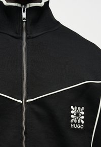 Μαύρο τζάκετ με φερμουάρ, λευκή διακόσμηση, ψηλό γιακά και κεντημένο λευκό ανθικό λογότυπο Hugo στο αριστερό στήθος.