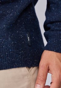 Pull en maille bleu marine avec texture marbrée, poignets côtelés et une petite étiquette de marque noire sur le côté, associé à un pantalon de couleur claire.