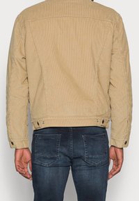 Giacca in velluto a coste color kaki con taglio dritto, dettagli cuciture visibili e bottoni a pressione regolabili sui polsini, abbinata a jeans scuri.