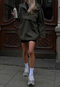 Personne marchant sur un trottoir en pierre, portant une veste vert olive, un short noir, des chaussettes blanches, des baskets grises, et tenant une bouteille d'eau argentée.