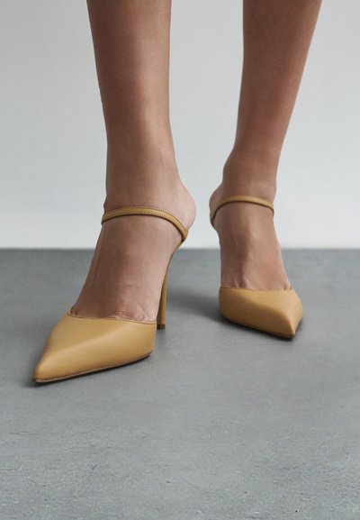 Žluté dámské nazouváky na vysokém podpatku s špičatou špičkou, tenkým páskem kolem kotníku, hladkou matnou texturou a minimalistickým designem na šedém povrchu.
