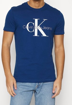 T-shirt en coton bleu avec un grand logo blanc "CK" et le texte "Calvin Klein Jeans" sur la poitrine, design classique à col rond.