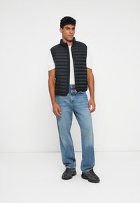 Black puffer vest with horizontal stitching, lightweight material, zippers, orange accent, worn over a white T-shirt and light blue jeans.