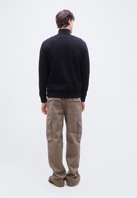 Άνδρας με μαύρο μακρυμάνικο μπλουζάκι και μπεζ παντελόνι φόρμας cargo στέκεται πλάτη, μπροστά σε απλό λευκό φόντο.